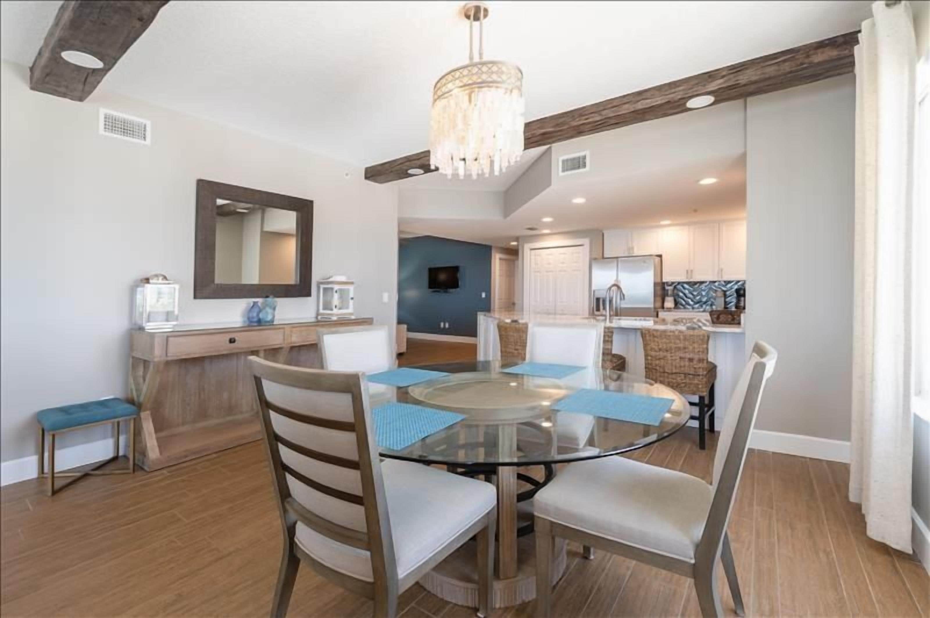 Un coin repas avec une table en verre, quatre chaises, un lustre, un banc en bois et, au fond, une cuisine.