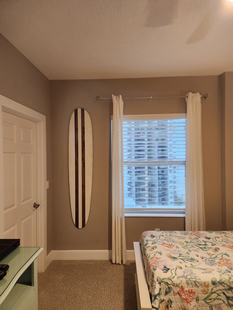A bedroom with a bed, a window with blinds, a surfboard on the wall, and a door.