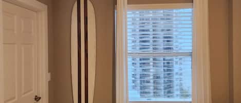 A bedroom with a bed, a window with blinds, a surfboard on the wall, and a door.