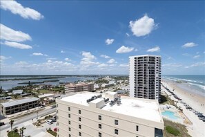 A coastal cityscape with a prominent high-rise hotel, a beach, and a clear blue sky. - Penthouse #1102 - Direct Oceanfront NE Corner  Luxury 3/3  Sanibel Condominium (Daytona Beach Shores)
