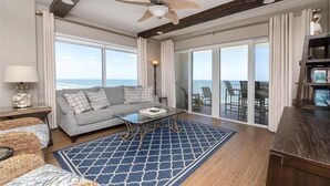 A spacious living room with a large window offering a view of the ocean, a ceiling fan, a sofa with cushions, a coffee table, and a bookshelf.