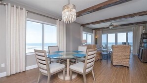 A dining area with a round glass table and four chairs, large windows with curtains, and a view of the ocean.
