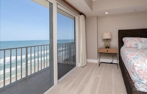 A hotel room with a large sliding glass door to the beach, a bed with floral bedding, a bedside table with a lamp, and a view of the ocean. - Penthouse #1102 - Direct Oceanfront NE Corner  Luxury 3/3  Sanibel Condominium (Daytona Beach Shores)