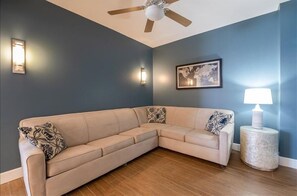 A beige sectional sofa with patterned pillows, a round side table with a lamp, and a ceiling fan in a room with dark blue walls and wooden flooring.