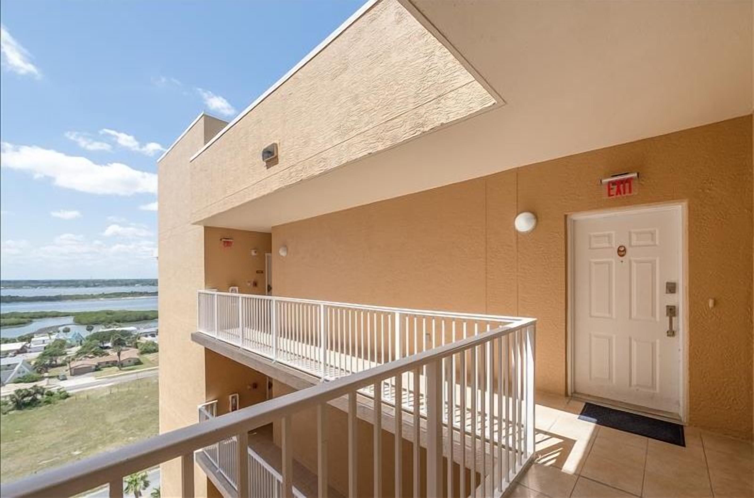 A balcony with a view of the sea and a building with a beige exterior, a white door, and an exit sign.