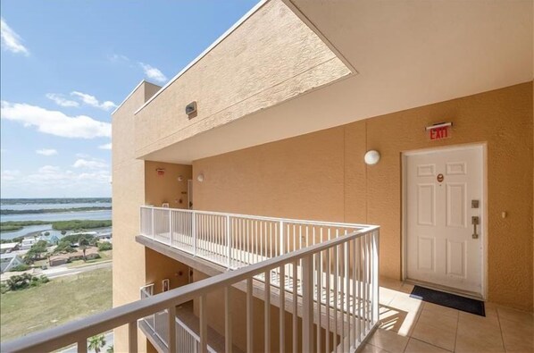 A balcony with a view of the sea and a building with a beige exterior, a white door, and an exit sign.