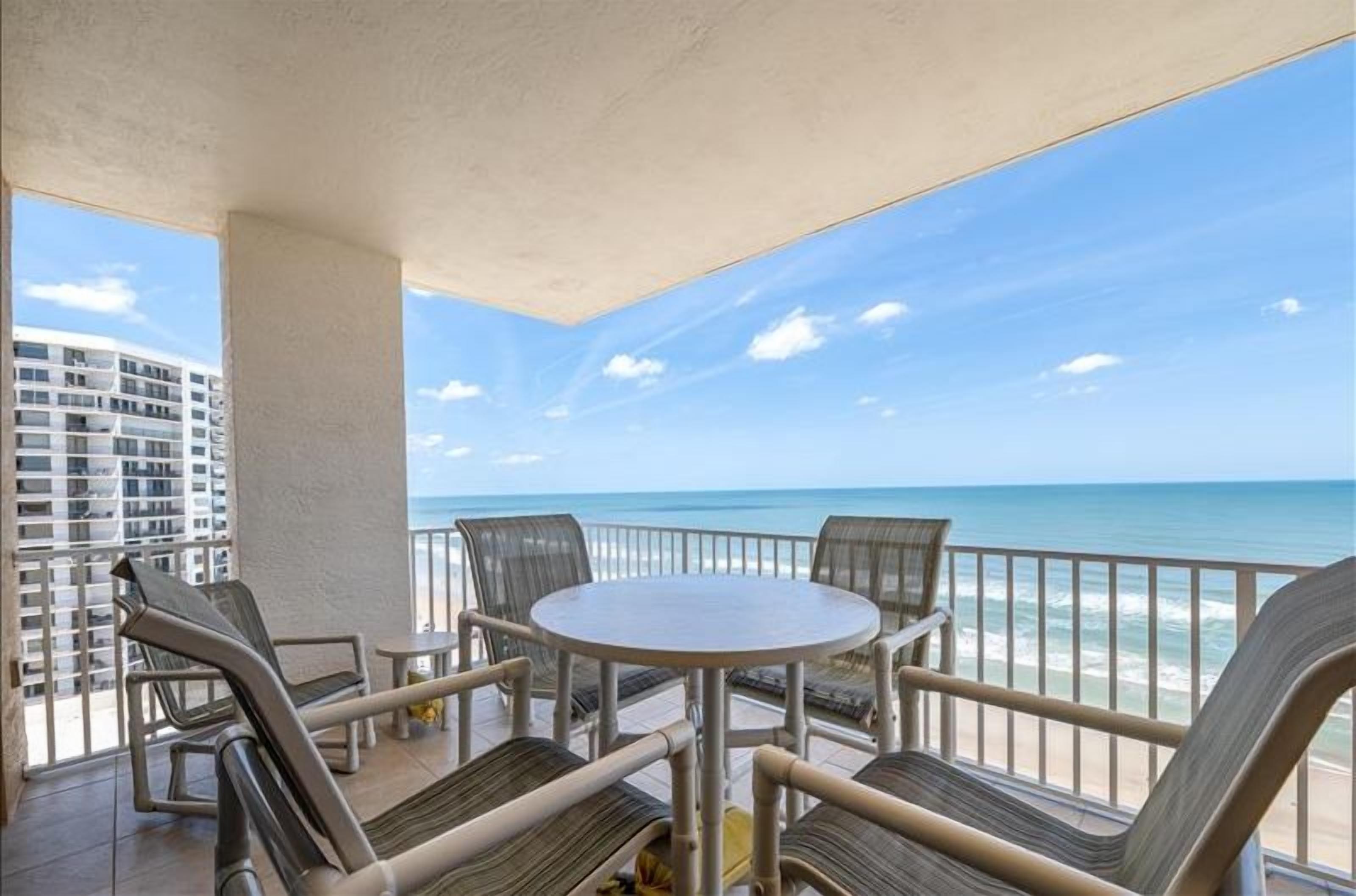 A balcony with a round table and chairs, overlooking a beach and a high-rise building.