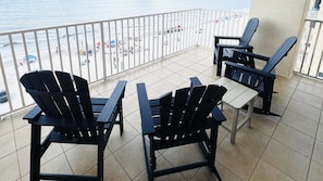 A balcony with three Adirondack chairs and a small table, overlooking a beach with umbrellas and people.