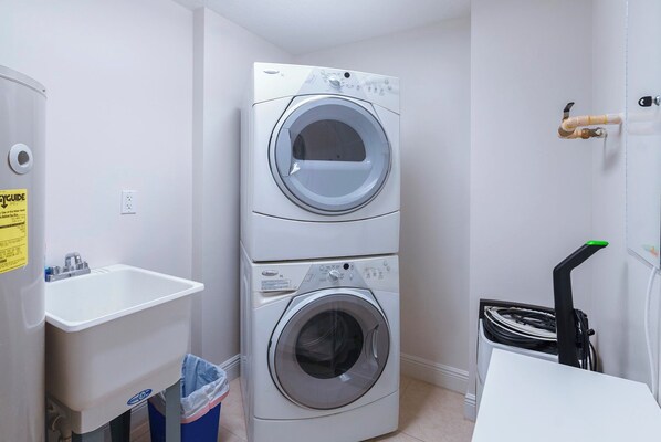 A laundry room with two stacked front-loading washing machines, a sink, and a trash bin.