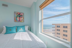 A bedroom with a bed, a window with a view of the beach and buildings, and a painting on the wall. - #705 Sanibel Direct Oceanfront SE Corner Unit 3/3 (Daytona Beach Shores)
