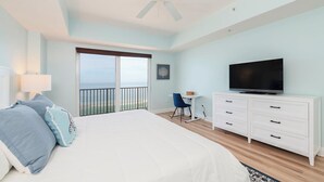 A bedroom with a large bed, a TV, a desk, and a balcony with a view of the sea.