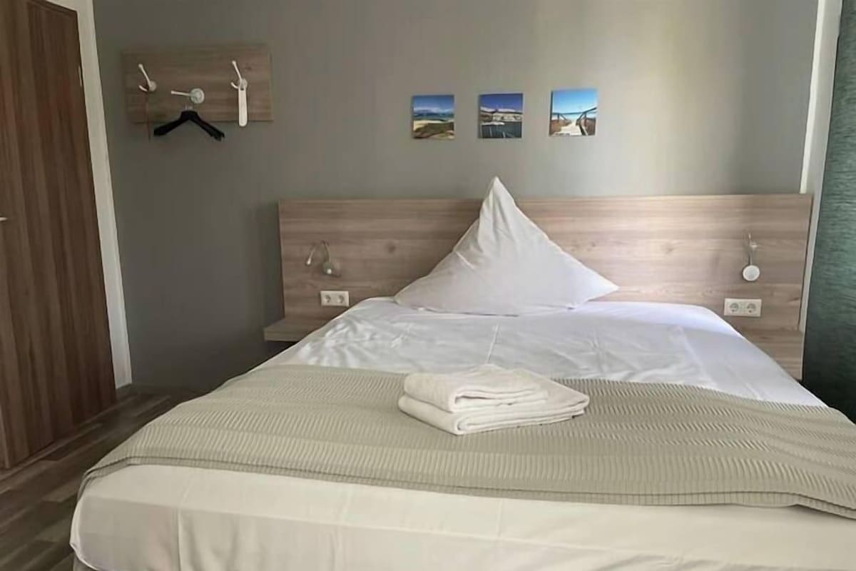 Un lit bien fait, avec une serviette blanche pliée, une tête de lit en bois et un porte-manteau mural avec des cintres.