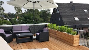 Une terrasse sur le toit, aménagée avec du mobilier d’extérieur, un jardinier en bois planté de végétaux et un grand parasol.
