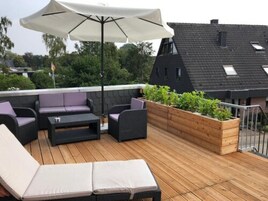 Um terraço no telhado com mobiliário externo, um cachepot de madeira com plantas e um grande guarda-sol.