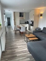 Uma sala de estar moderna com um sofá cinzento, uma mesa de centro de madeira e um espaço de jantar com uma mesa e cadeiras brancas.