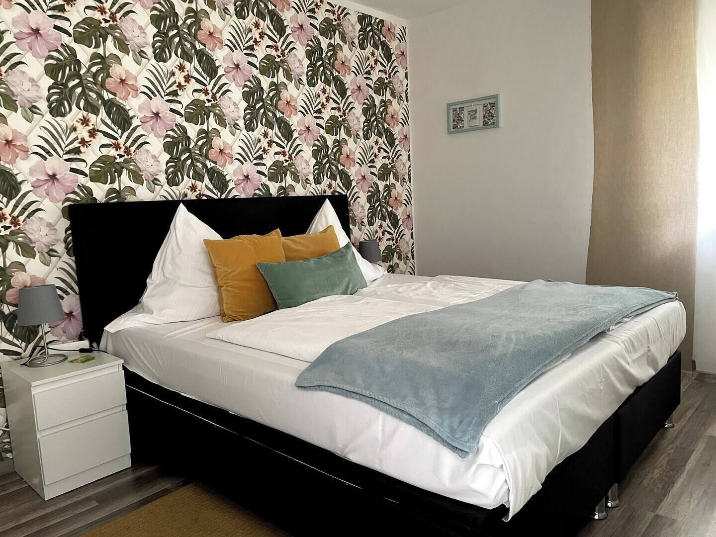 A bedroom with a floral wallpaper, a black headboard, white bedding, and a bedside table with a lamp.