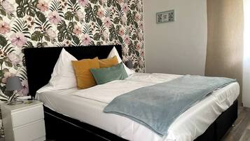 A bedroom with a floral wallpaper, a black headboard, white bedding, and a bedside table with a lamp.