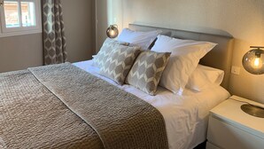 A neatly made bed with patterned pillows and a textured blanket, a bedside lamp, and a window with curtains. - Cathedral Aparthotel historic center cathedral view Superior (Albi)