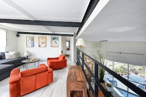 A modern living room with a sofa, chairs, a wooden bench, and a table. There are artworks on the wall and a view of the outside through large windows.