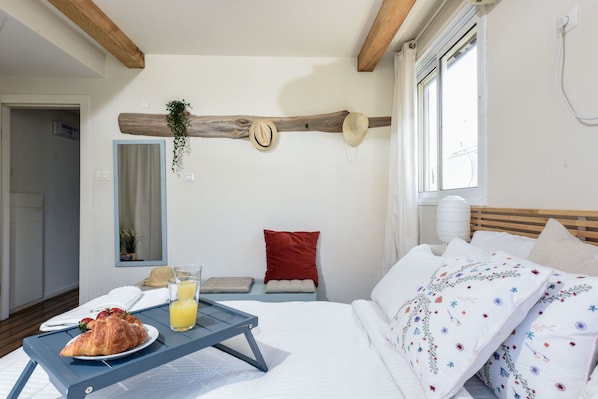 A hotel room with a bed, a breakfast tray with croissant and orange juice, a red pillow, and a wooden beam ceiling.