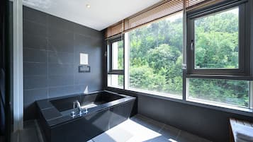 A modern bathroom with a large black bathtub, large windows offering a view of greenery, and a wooden bench.