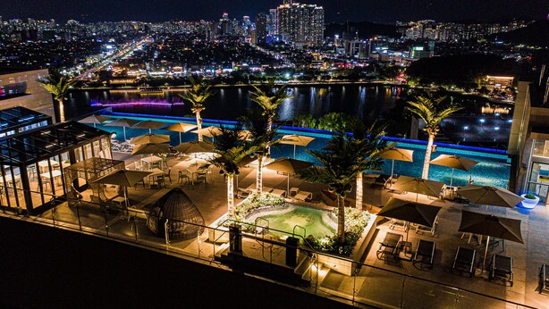 A rooftop pool area with lounge chairs and umbrellas, overlooking a cityscape at night.