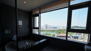 A modern bathroom with a large bathtub, a sink, and a window offering a city view.