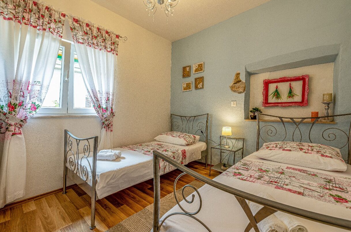 A bedroom with two single beds, a window with floral curtains, a chandelier, and wall-mounted picture frames.
