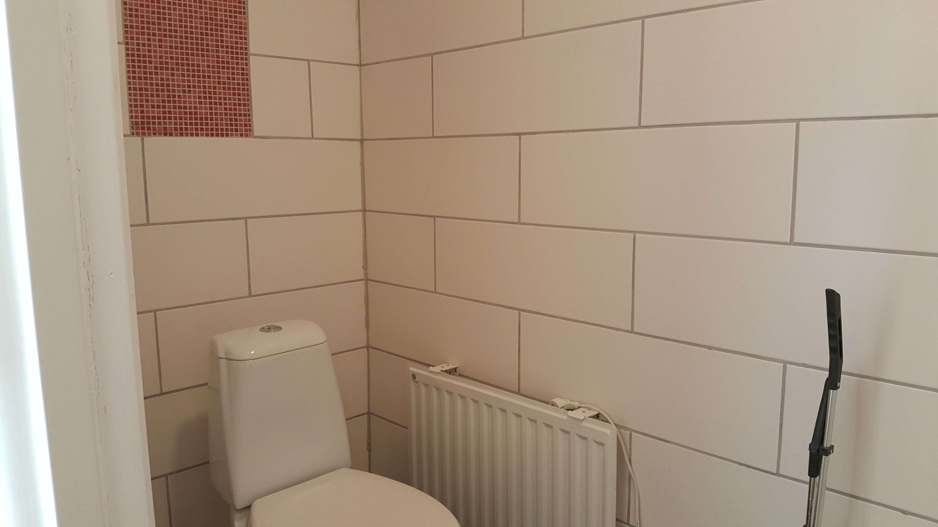 A small bathroom with white tiles, a toilet, a radiator, and a black cleaning tool leaning against the wall.