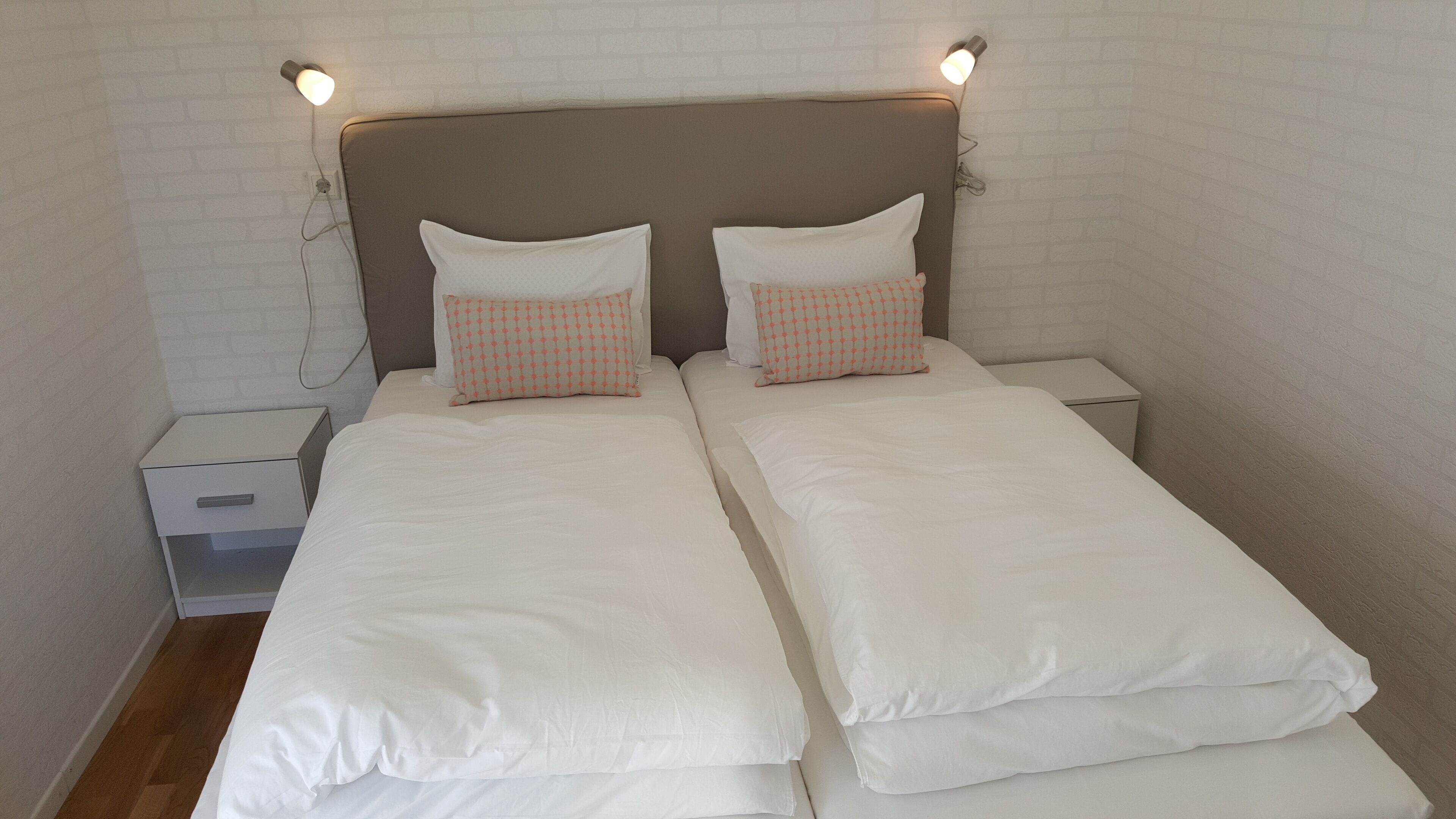 A double bed with white bedding and two patterned pillows, a nightstand, and a wall-mounted light fixture.