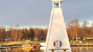 Un faro con un reloj en su lateral, un muelle con barcos, y casas al fondo.