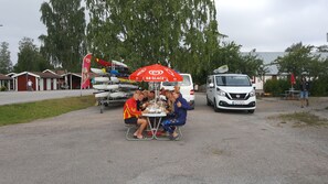 Un grupo de personas sentadas a una mesa bajo un paraguas GB GLACE, con una fila de kayaks al fondo.