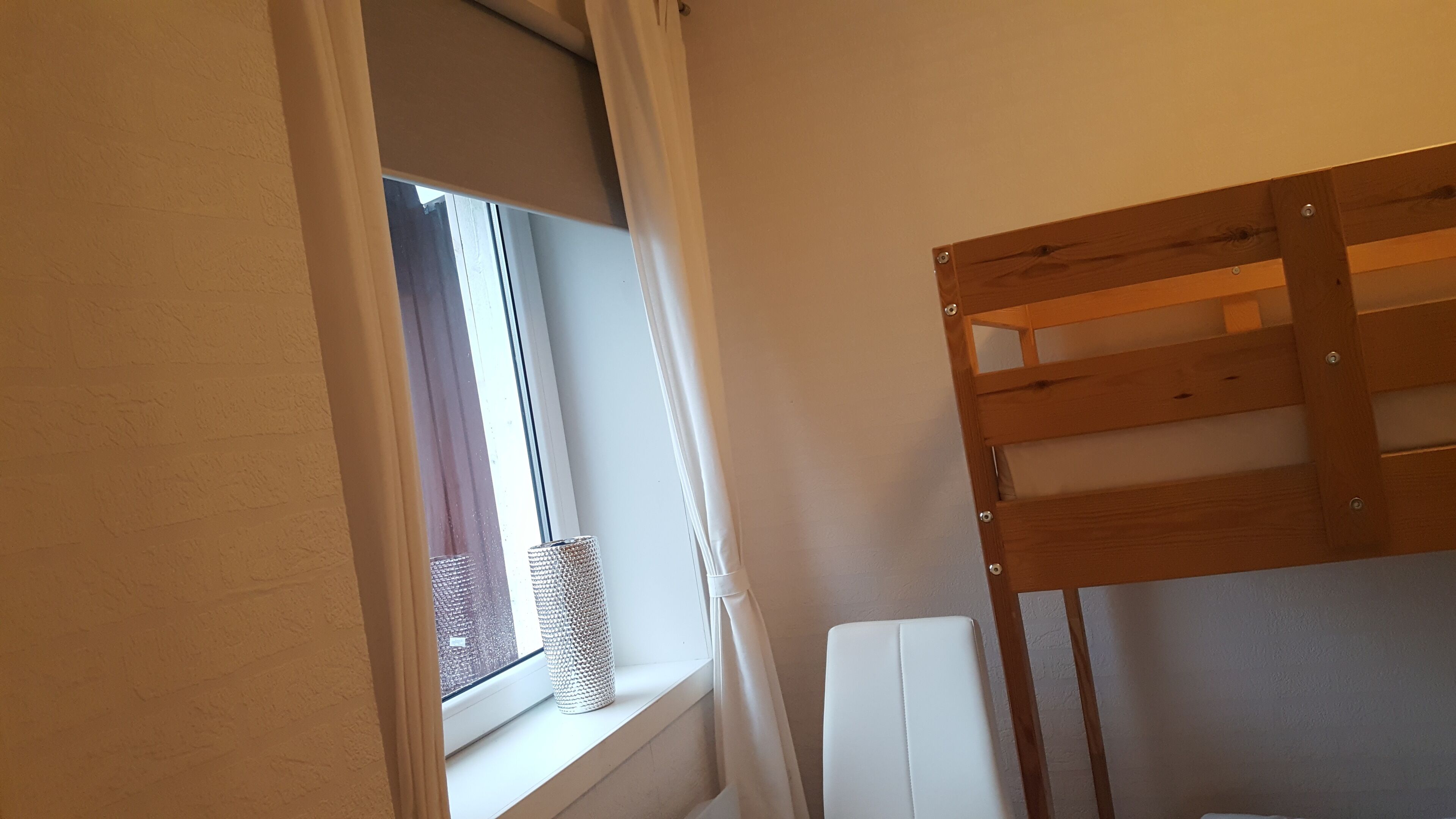 Una cama litera con estructura de madera y colchón blanco. Hay una ventana con persiana blanca que la cubre parcialmente, y una pequeña cesta en el alféizar.