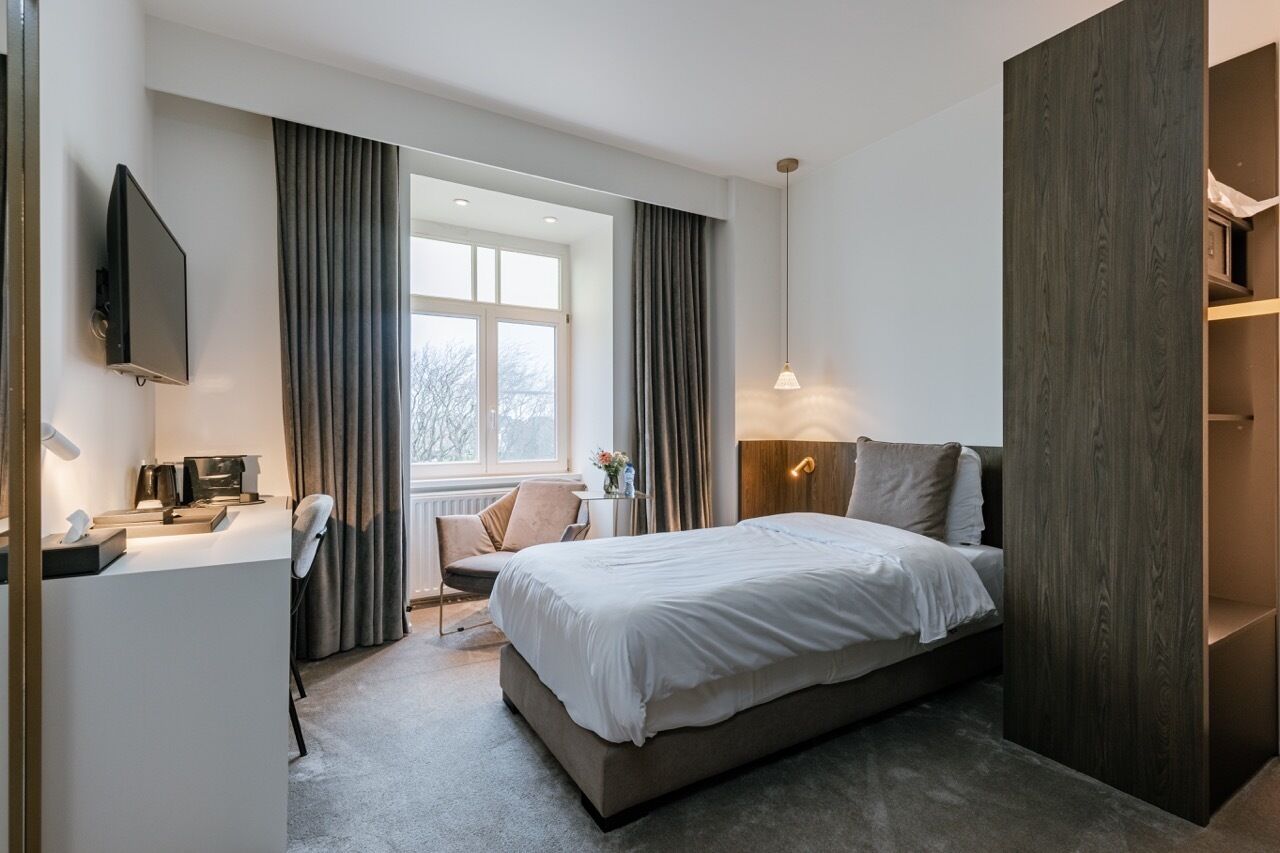 Moderni hotellihuone, jossa on suuri sänky, työpöytä tietokoneella, televisio ja ikkuna, jossa on verhot.