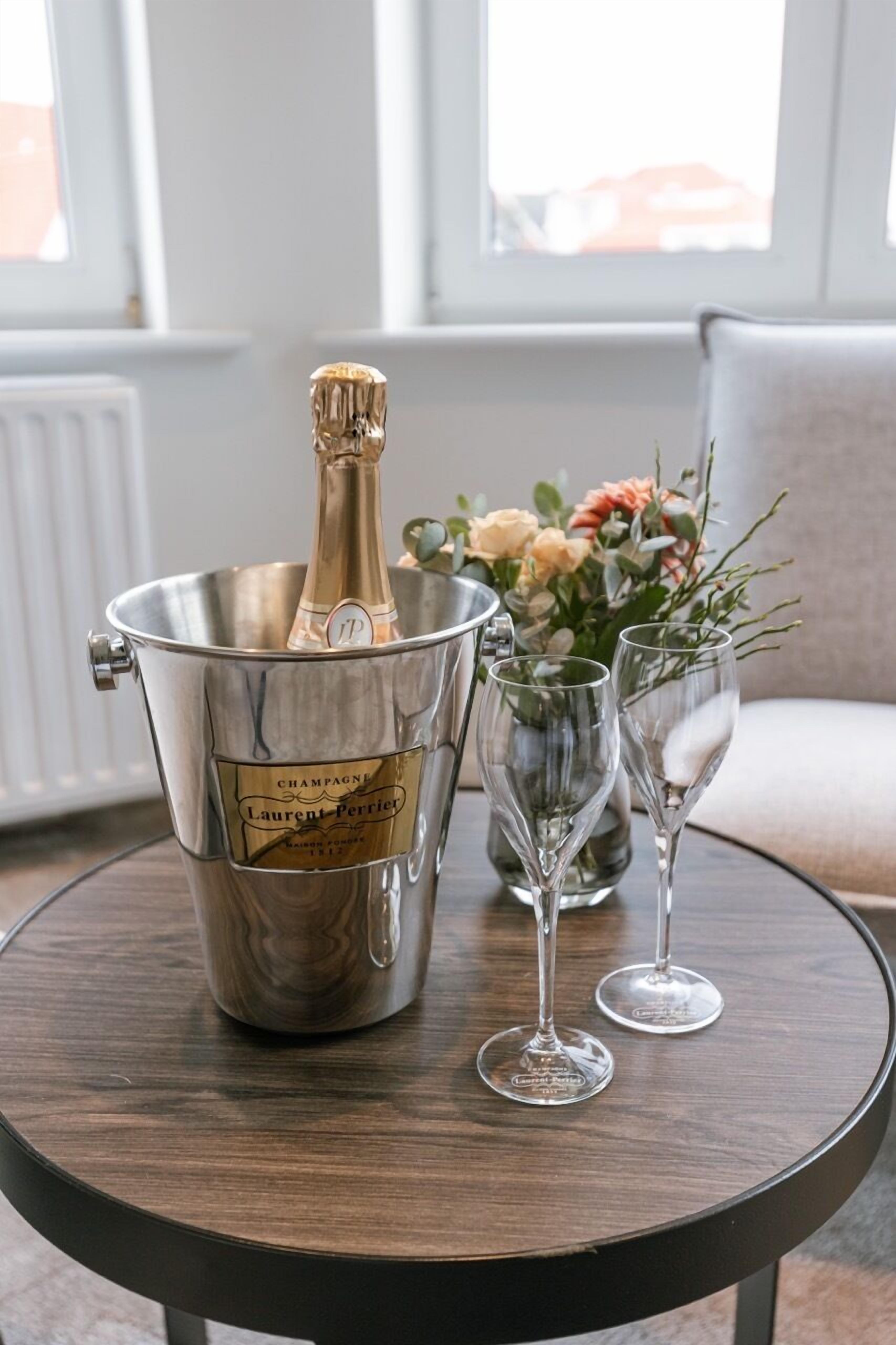Un balde metálico para champán con una botella, dos copas de vino vacías y un florero con flores sobre una mesa redonda de madera.