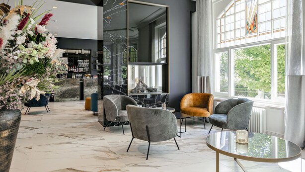 A modern interior with a marble floor, grey upholstered chairs, a glass table, and a large mirror. - Hotel Astoria De Haan (De Haan)