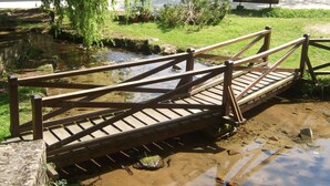A wooden bridge over a muddy stream with a stone wall on one side and a building with a red roof in the background. - Camino Real de Selores (Valle de Cabuerniga)