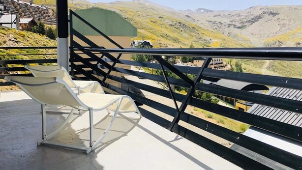 Un balcon avec une chaise blanche, une rambarde en métal et une vue sur un paysage montagneux, avec des bâtiments en arrière-plan.