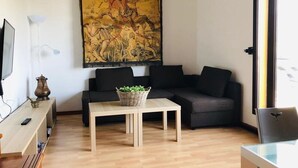 Un salon moderne avec un plancher en bois, un canapé noir, une petite table avec un panier, un téléviseur posé sur un meuble et un grand tableau accroché au mur.