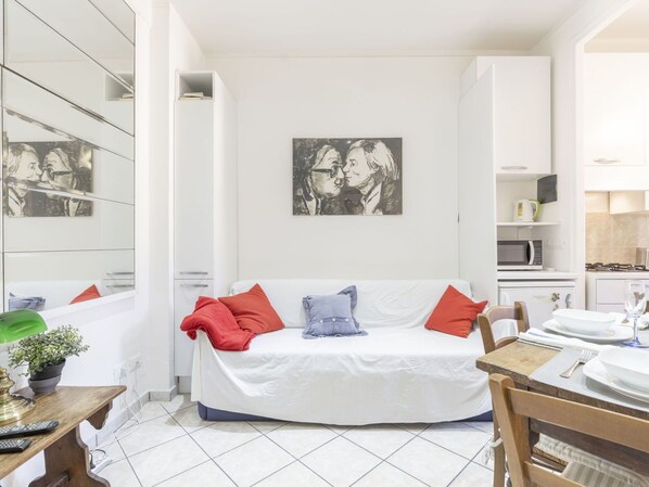 A modern living room with a white sofa, red and blue cushions, a painting on the wall, and a dining area with a wooden table and chairs. - Cosy apartment for 4 guests with WIFI, TV, balcony and pets allowed (Sanremo)