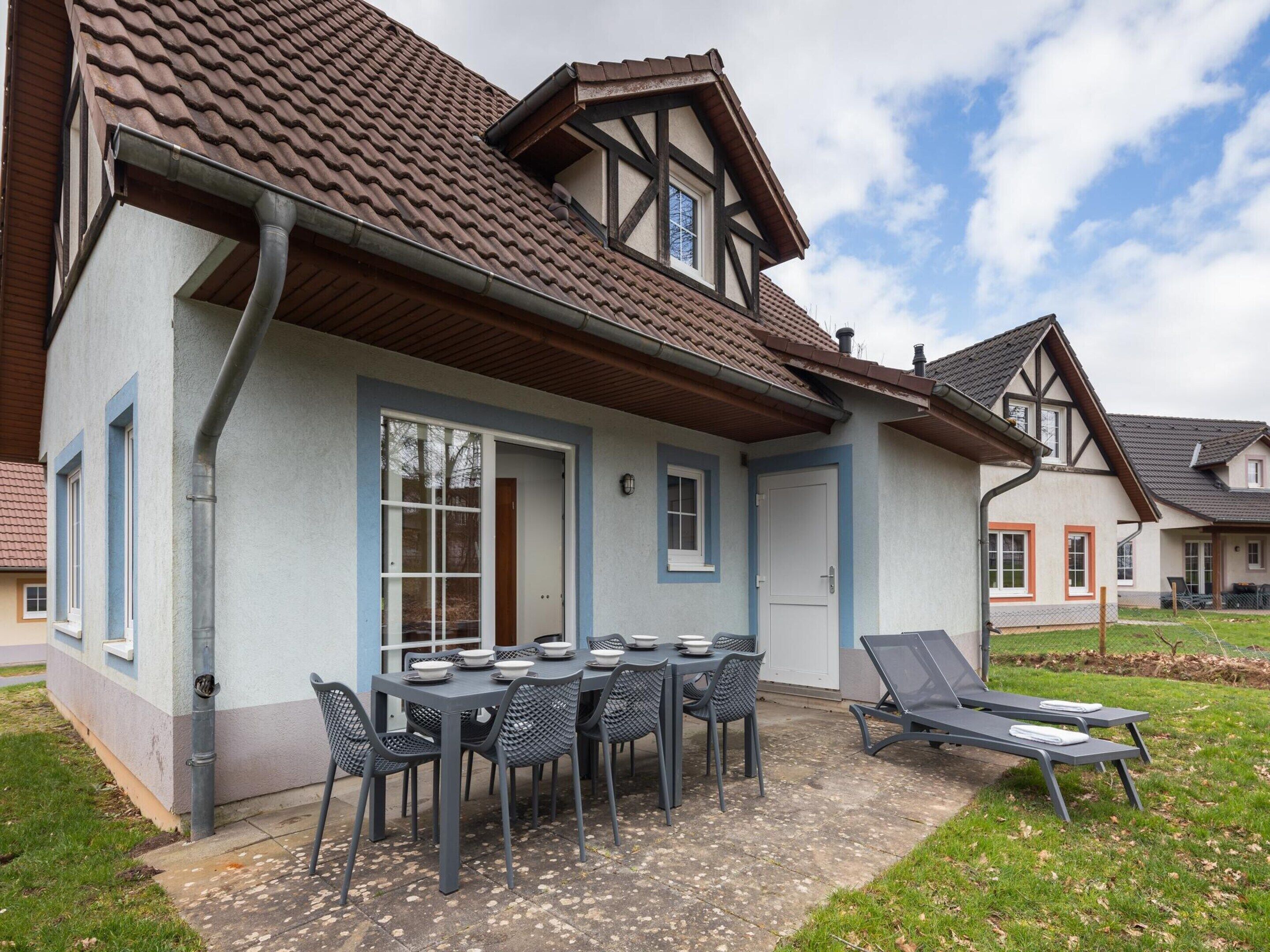 A house with a tiled roof, a dining area with a table and chairs, and a small outdoor seating area with lounge chairs.