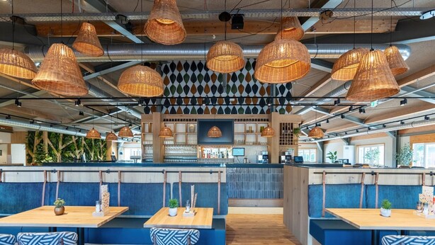 Un restaurante moderno con mesas de madera, asientos tapizados en azul y lámparas colgantes de mimbre.