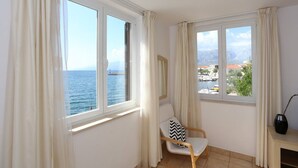 A room with two windows overlooking the sea and a view of buildings. There is a chair with a black and white pillow and a lamp on a side table. - Bech Apartment - 4 Persons (Posedarje)