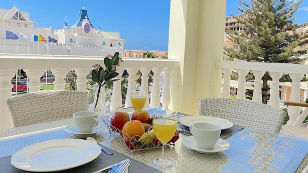 Ein Balkon mit Frühstückstisch, dekoriert mit Früchten und einem Glas Orangensaft, mit Blick auf ein Gebäude mit blauem Dach und Fahnen.