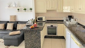 Eine kompakte Küche mit Mikrowelle, Backofen und Spüle. Ein schwarzes Sofa mit gemusterten Kissen und ein kleiner Couchtisch stehen im Raum. Die Küche verfügt über helle Schränke und eine Granitarbeitsplatte.