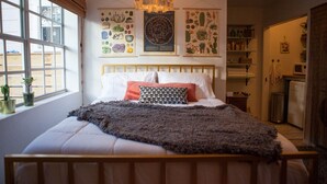 Un dormitorio con cama de madera, ropa de cama blanca, un edredón gris y una lámpara colgante. Hay carteles botánicos en la pared y una ventana con plantas en el alféizar.
