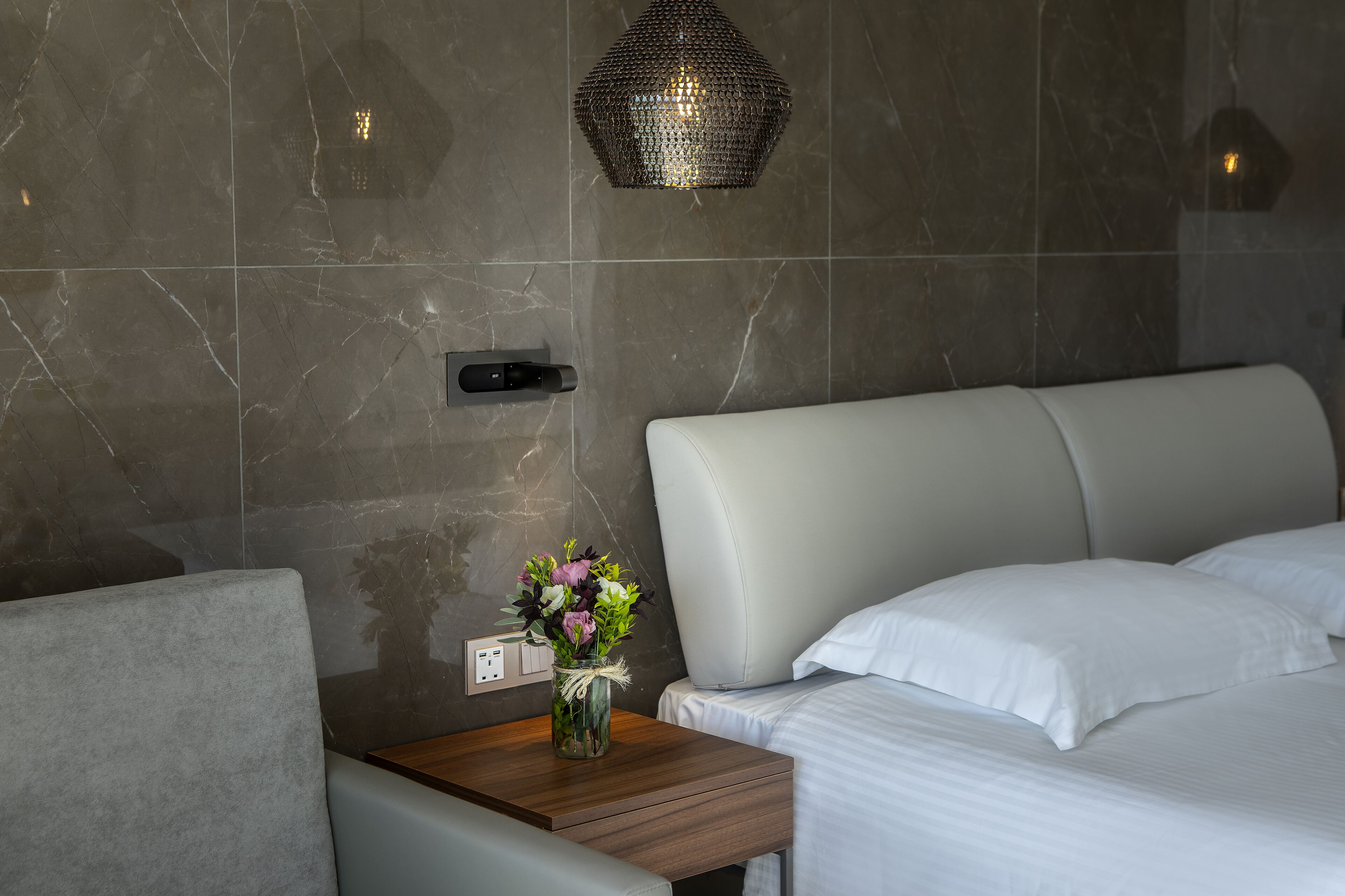 Una habitación de hotel moderna con pared de mármol, una mesita de noche con un jarrón de flores, una cama con ropa de cama blanca e iluminación de techo con textura.
