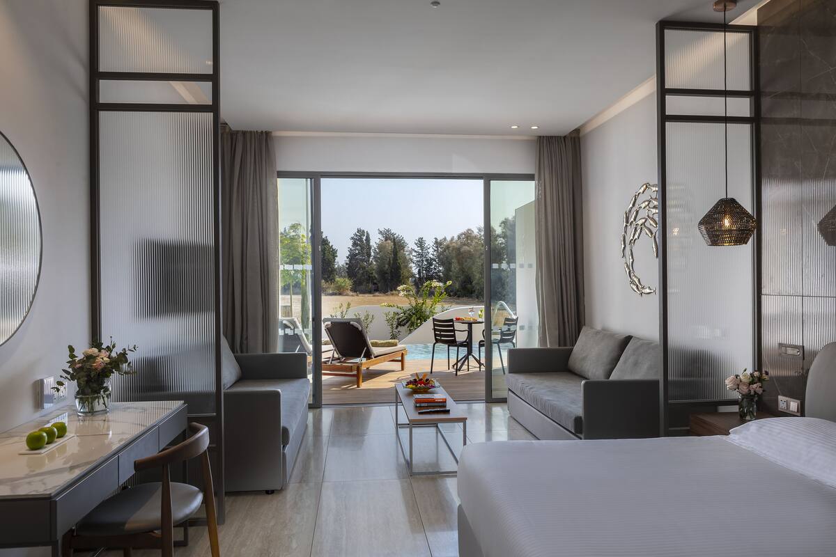 Une chambre d’hôtel moderne dotée d’un grand lit, d’un bureau avec une chaise, d’un canapé, d’un coin repas avec une table et des chaises, et d’un balcon offrant une vue sur l’extérieur.