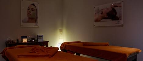 Een kamer met twee massagetafels, een bureau met lamp en wanddecoraties in lijsten.