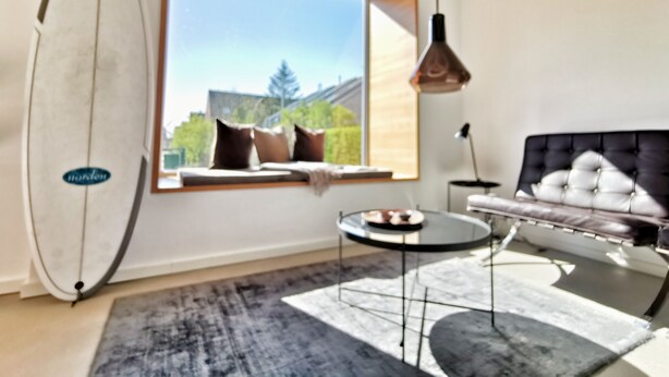 A modern room with a large window, a black upholstered sofa, a round coffee table, and a surfboard leaning against the wall. - Modern small vacation home with southwest-facing garden (shed XXXI) (Norderney)